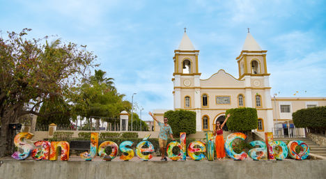 Vibrant oversized "San José del Cabo" sign with two people posing in front of a white twin-towered church, palm trees and bright blue sky in San José del Cabo, Mexico.
