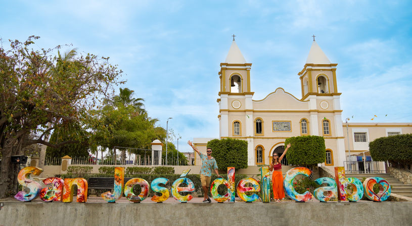 Vibrant oversized "San José del Cabo" sign with two people posing in front of a white twin-towered church, palm trees and bright blue sky in San José del Cabo, Mexico.