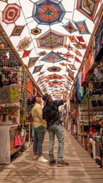 Two shoppers gaze up at a vibrant canopy of colorful woven geometric hangings above a bustling artisan market aisle lined with handmade textiles, bags, and crafts.