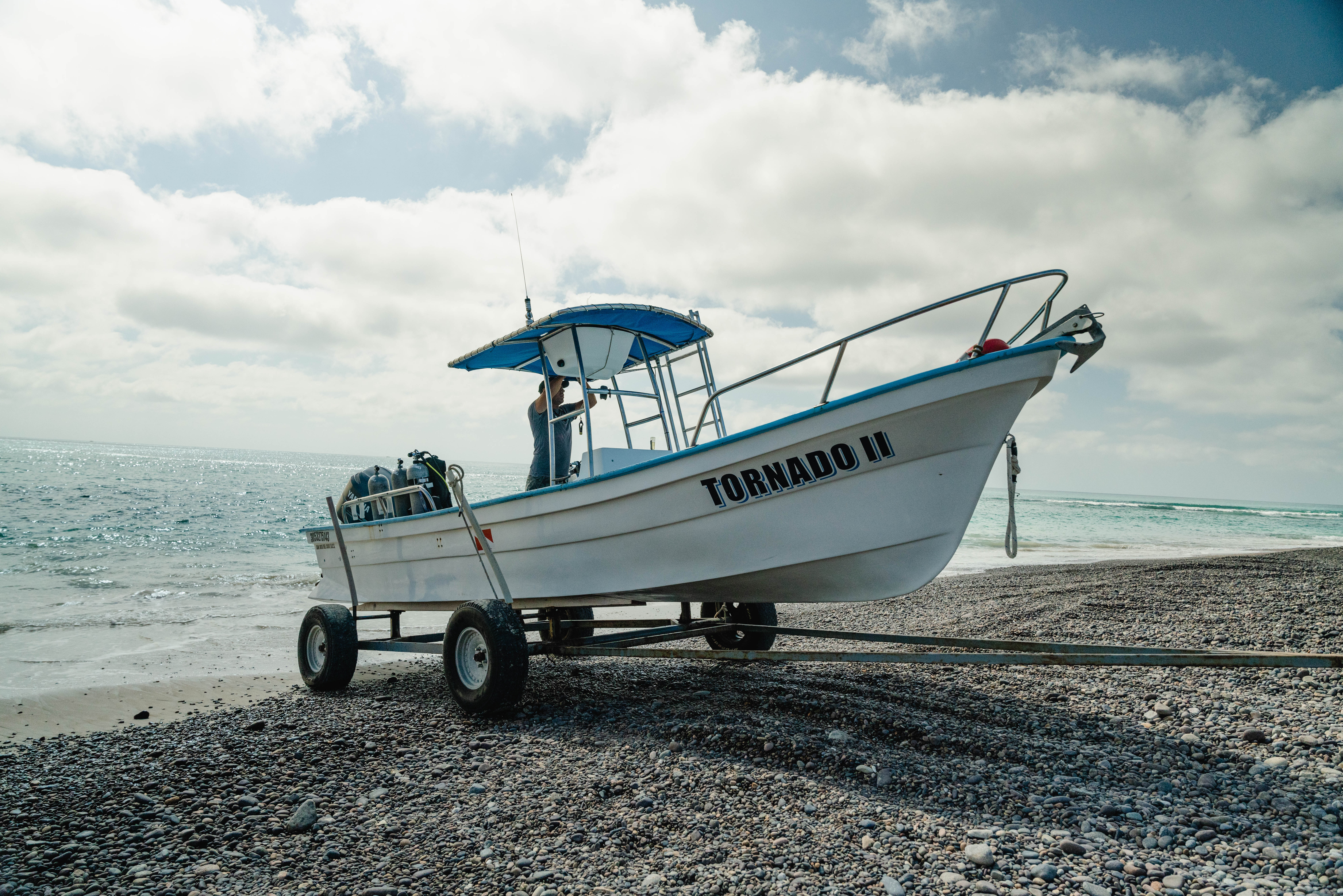 White-and-blue motorboat on a wheeled trailer parked on a pebble beach, scuba tanks visible, calm turquoise ocean and cloudy sky