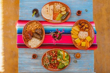 Overhead view of a vibrant Mexican-style spread on a blue wooden table with a colorful serape runner — terracotta plates of rice, beans, tortillas, roasted potatoes, corn, salad, chips, drinks, and a small pink-flowered centerpiece.