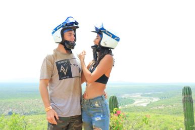 Two off-road riders in blue-and-white helmets smile at each other on a sunlit desert overlook dotted with tall cacti and wildflowers, a winding river valley stretching into the distance.