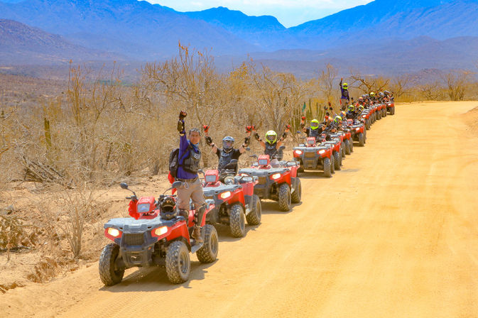 Group ATV tour on a dusty desert trail with red ATVs lined up on a dirt road, riders cheering and raising hands amid arid scrub and a blue mountain backdrop
