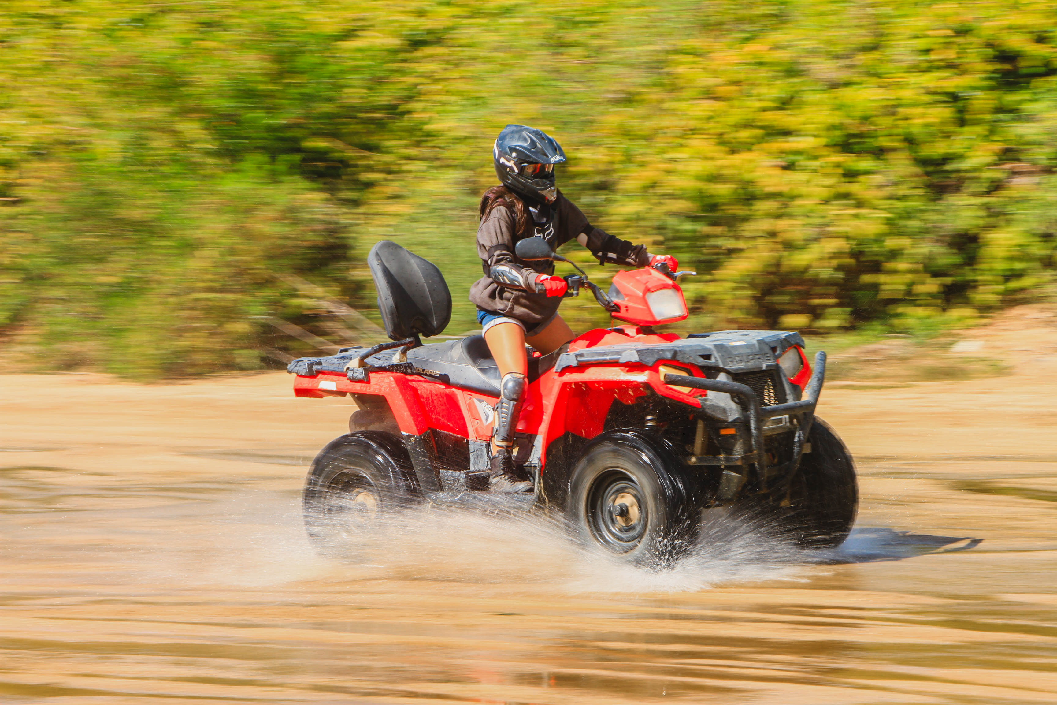 Person in helmet and knee pads riding a red ATV, splashing through shallow water on a sandy off-road trail with green foliage in the background.