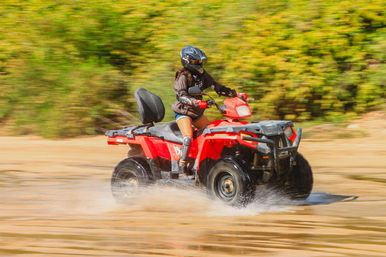Person in helmet and knee pads riding a red ATV, splashing through shallow water on a sandy off-road trail with green foliage in the background.