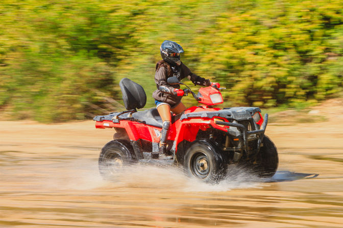 Person in helmet and knee pads riding a red ATV, splashing through shallow water on a sandy off-road trail with green foliage in the background.