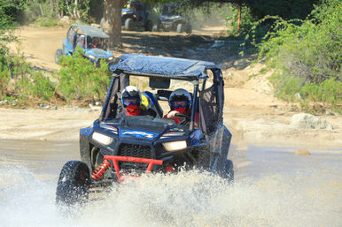 Two riders in a blue off-road UTV (side-by-side) splashing through shallow water on a dusty trail, wearing helmets and goggles with trees and other UTVs in the background.