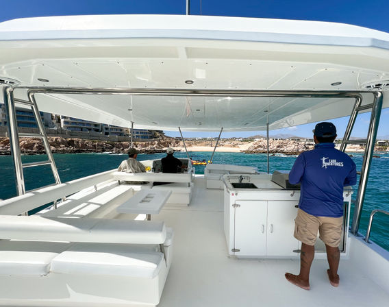 Spacious white boat deck with cushioned seating and shaded canopy, crew member in a blue shirt standing by a small kitchenette, two passengers seated toward the bow, turquoise ocean and rocky beachfront with low-rise buildings under a clear blue sky