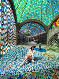 Person in a straw hat and white outfit seated on a vibrant blue mosaic floor beneath a multicolored stained-glass dome and arched brick tile walls in a bright indoor atrium.