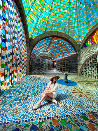 Person in a straw hat and white outfit seated on a vibrant blue mosaic floor beneath a multicolored stained-glass dome and arched brick tile walls in a bright indoor atrium.