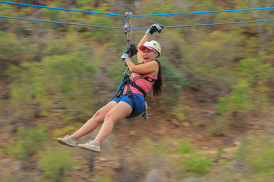 Woman ziplining over a green hillside, wearing helmet, gloves and harness, smiling during an outdoor adventure