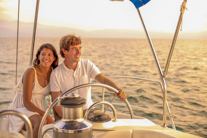 Smiling couple on a sailboat at golden sunset, man at the helm steering while woman leans close, calm ocean and distant horizon.
