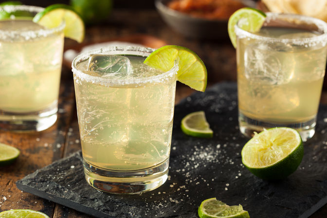 Zesty salt-rimmed lime margarita on the rocks in a clear glass with lime wedge, ice cubes and halved limes on a slate board in a rustic setting