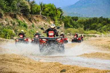 Group of riders on red ATVs splashing through a shallow stream on a sandy off-road trail with green scrub and distant mountains — outdoor off-road adventure.