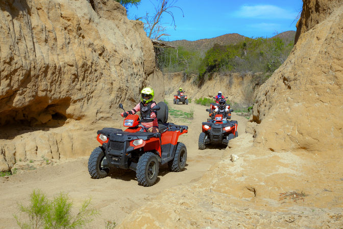 Three helmeted riders on red ATVs navigating a narrow sandy desert canyon trail between eroded tan rock walls under a bright blue sky — off-road ATV adventure.