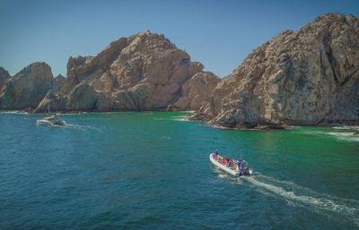 Motorized tour boat and sailboat cruising turquoise ocean near dramatic sunlit rocky sea cliffs under a clear blue sky
