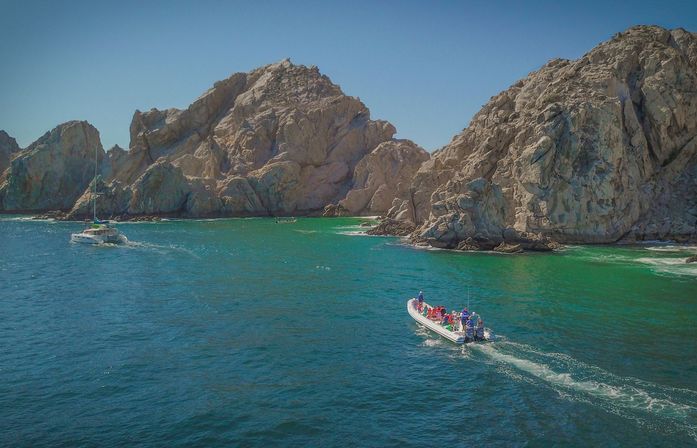 Motorized tour boat and sailboat cruising turquoise ocean near dramatic sunlit rocky sea cliffs under a clear blue sky