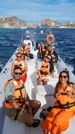 Happy group in orange life jackets on a RIB boat enjoying a sunny boat tour on deep blue water toward a coastal resort with hillside buildings