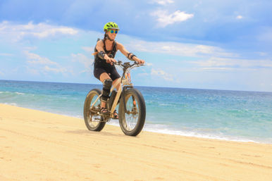 Cyclist wearing a green helmet and sunglasses riding a fat-tire bike along a sandy ocean beach with turquoise water and a partly cloudy blue sky