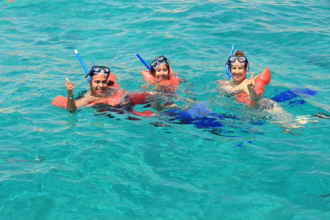 Three smiling snorkelers in orange life jackets and blue snorkels floating in clear turquoise tropical water, giving thumbs-up and peace sign