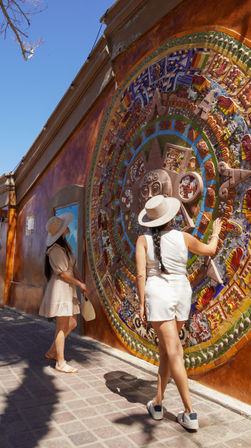 Two women in sun hats admire a large, colorful Aztec-style mosaic sun mural on an orange stucco wall in a sunny pedestrian plaza.