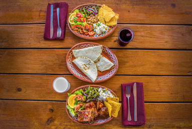 Three terracotta plates of Mexican-style dishes on a rustic wooden table: folded tortillas, enchiladas, rice, beans, fresh salad, tortilla chips, dark and light drinks, and cutlery on burgundy napkins.