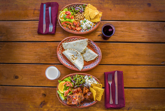 Three terracotta plates of Mexican-style dishes on a rustic wooden table: folded tortillas, enchiladas, rice, beans, fresh salad, tortilla chips, dark and light drinks, and cutlery on burgundy napkins.