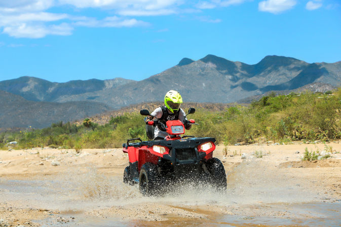 ATV rider in a neon yellow helmet on a red all-terrain vehicle splashing through a shallow desert wash with scrubby vegetation and rugged mountains under a bright blue sky