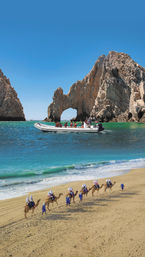 Camel caravan led by guides in blue robes along a golden beach, tourists on camels with white head coverings, turquoise sea with a speedboat near the iconic Cabo San Lucas rock arch (El Arco) under a clear blue sky.