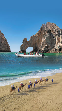 Camel caravan led by guides in blue robes along a golden beach, tourists on camels with white head coverings, turquoise sea with a speedboat near the iconic Cabo San Lucas rock arch (El Arco) under a clear blue sky.