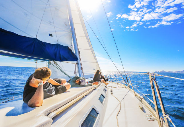 Sun-drenched sailboat deck with people lounging under full white sails and blue canvas, sparkling open ocean and distant coastline