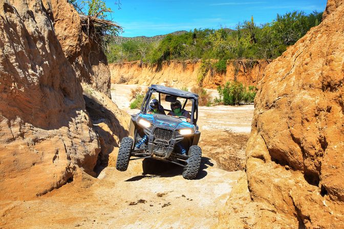 Two helmeted riders in a blue off-road UTV navigating a narrow sandy desert canyon trail between orange sandstone walls under a bright blue sky