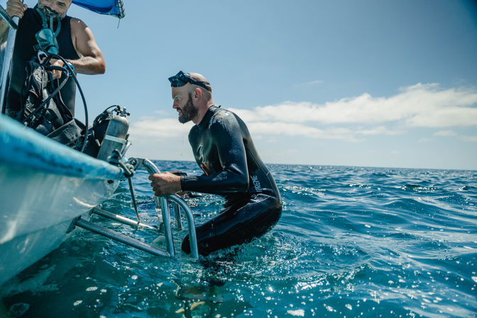 Scuba diver in a black wetsuit climbing a boat ladder from the open ocean while a crew member readies scuba tanks under a sunny blue sky.