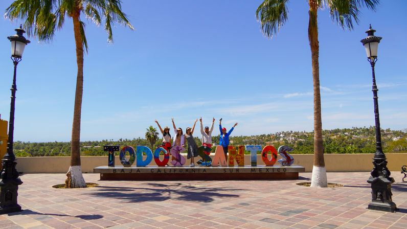 Vibrant 'Todos Santos' letters on a sunny Baja California Sur promenade, palm trees and vintage lampposts framing a group of people posing against a clear blue sky and coastal landscape in Mexico.