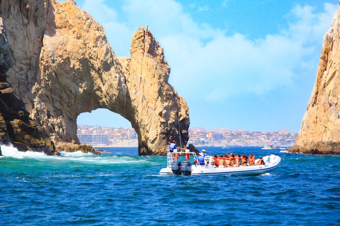 Speedboat of tourists in life jackets cruising turquoise sea beneath the iconic El Arco rock arch at Cabo San Lucas, Mexico, with rugged cliffs and a distant coastal skyline.