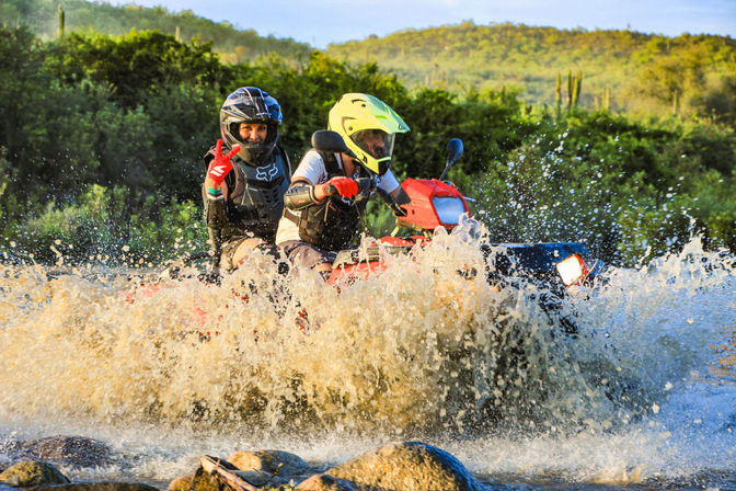 Two off-road ATV riders in helmets and protective gear splashing through a shallow river at golden hour, with green scrubby hills and cacti in the background — outdoor trail adventure.