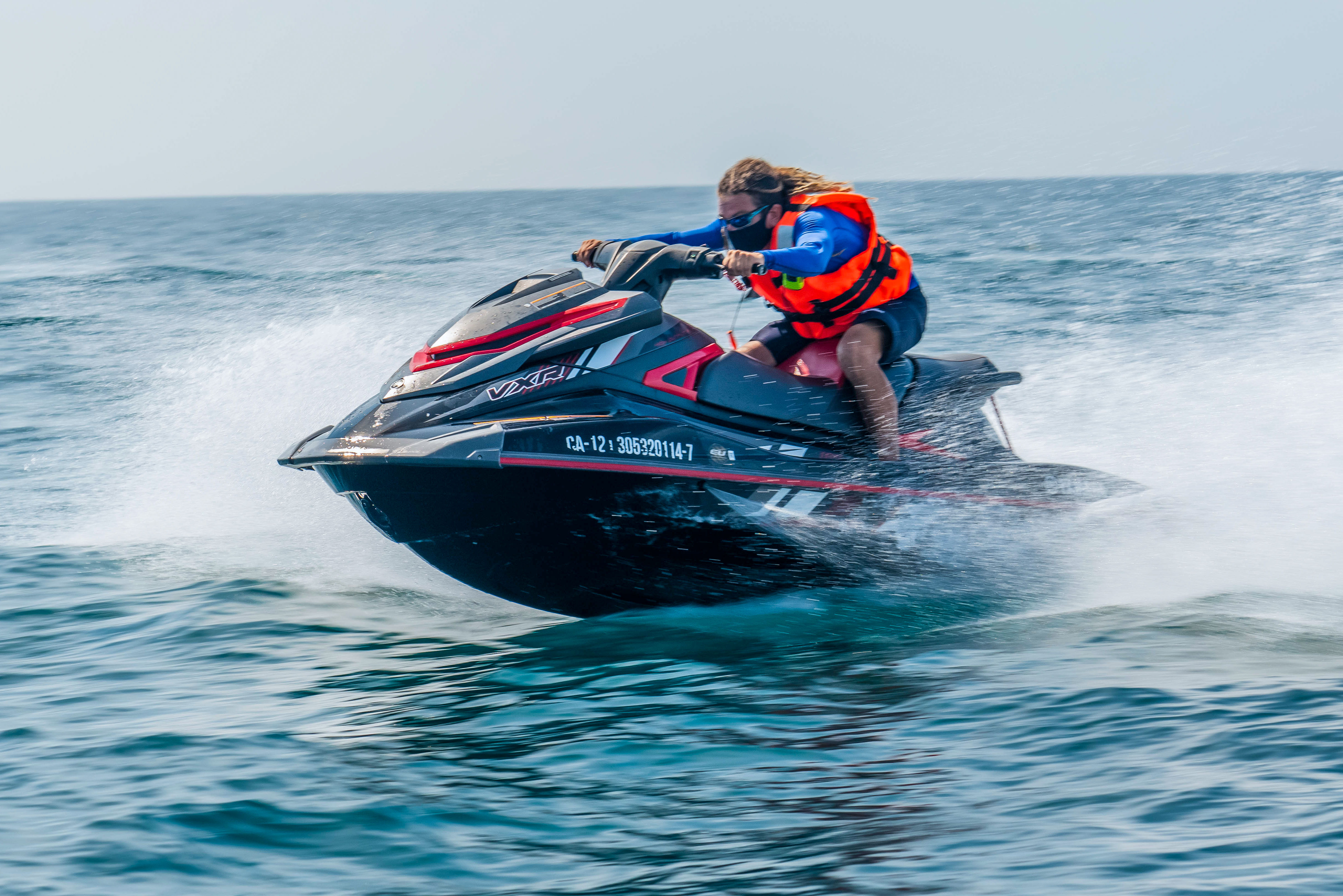 Person in an orange life jacket and blue sleeves riding a black-and-red jet ski across choppy blue ocean water, kicking up spray and speeding over waves