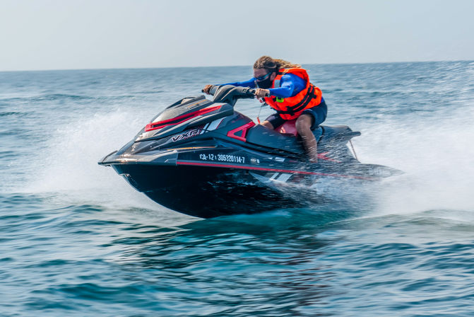 Person in an orange life jacket and blue sleeves riding a black-and-red jet ski across choppy blue ocean water, kicking up spray and speeding over waves