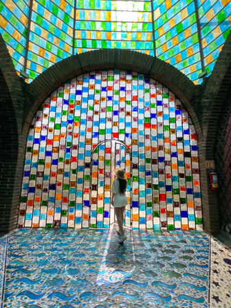 Person wearing a sunhat stands before a large arched stained-glass wall of colorful square panes, sunlight casting vibrant mosaics onto a blue wave-pattern tiled floor in a bright interior.