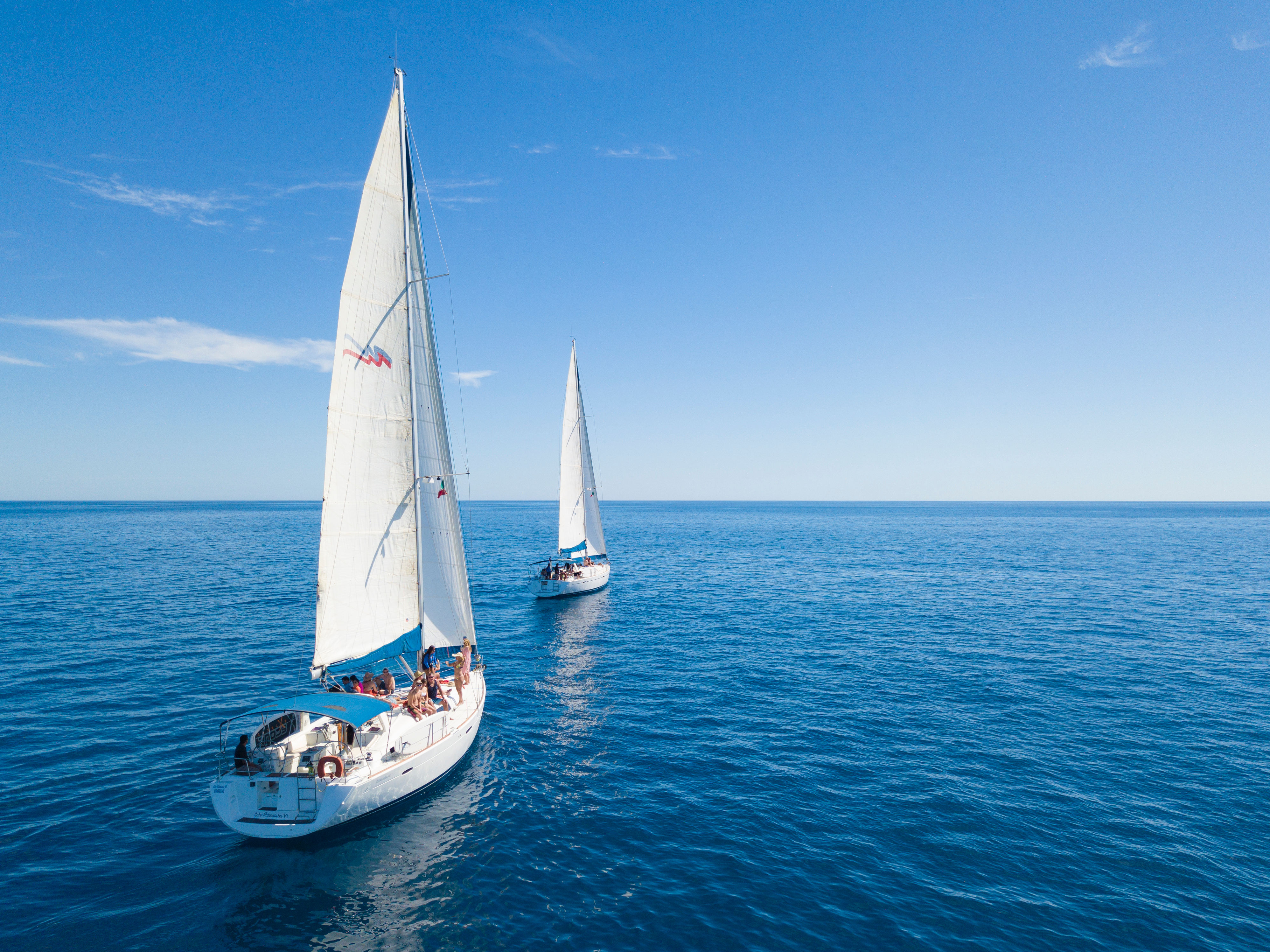 Aerial view of two white sailboats gliding across calm deep-blue sea under a clear blue sky, passengers relaxing on deck.