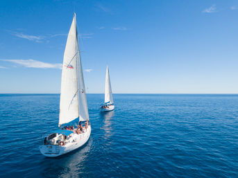 Aerial view of two white sailboats gliding across calm deep-blue sea under a clear blue sky, passengers relaxing on deck.