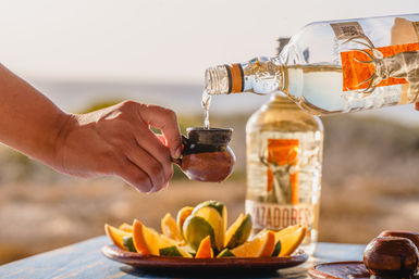 Hand pouring clear tequila from a bottle into a small rustic clay cup next to a plate of lime and orange wedges on a table with a sunlit seaside golden-hour background