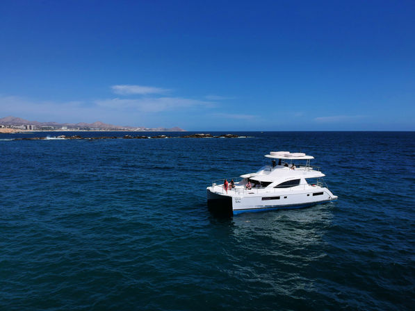 White luxury catamaran with passengers on deck cruising deep-blue ocean near a rocky coastline and distant dry hills under a clear blue sky