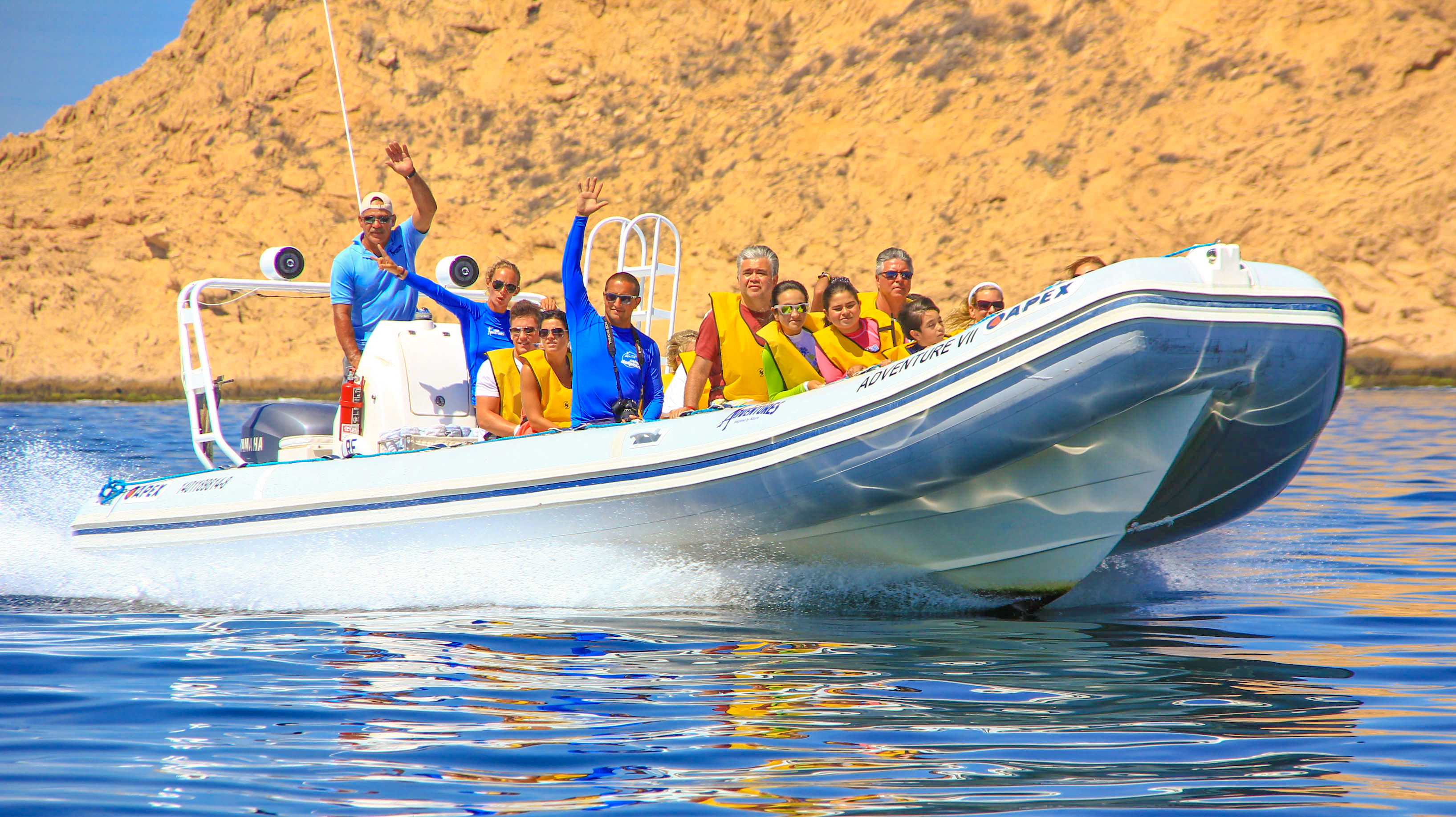 Smiling group wearing yellow life jackets on a white inflatable speedboat speeding across clear blue water past sandy coastal cliffs on a sunny day