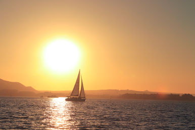 Sailboat silhouetted on calm ocean during a golden sunset, sun reflecting a bright path across the water with distant coastal hills.