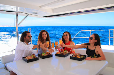Four adults clinking mimosa glasses over boxed brunch on a sunlit yacht deck with vivid blue ocean and clear sky, luxury ocean cruise vibe.