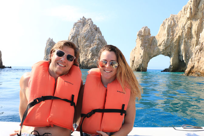 Two smiling boaters in orange life jackets and sunglasses posing on a boat in front of the El Arco rock formation at Cabo San Lucas, Mexico, with bright blue sea and sky