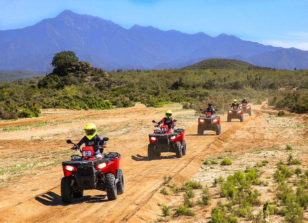 Five riders in neon helmets on red ATVs ride single-file along a dusty off-road trail through arid scrubland with low green bushes and a blue mountain backdrop under clear skies.