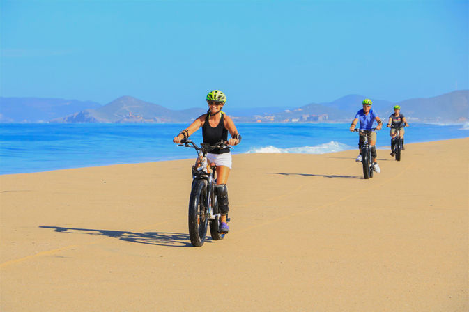 Three cyclists wearing helmets ride fat-tire bikes along a sunny sandy beach beside the blue ocean with distant coastal hills — upbeat seaside bike ride scene.