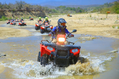 Helmeted rider on a red ATV splashing through a shallow desert riverbed, followed by a line of ATVs on a sandy off-road canyon trail.